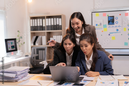 Three excited asian businesswomen are celebrating success while working together on a laptop in a modern office, surrounded by paperwork and a whiteboard with charts