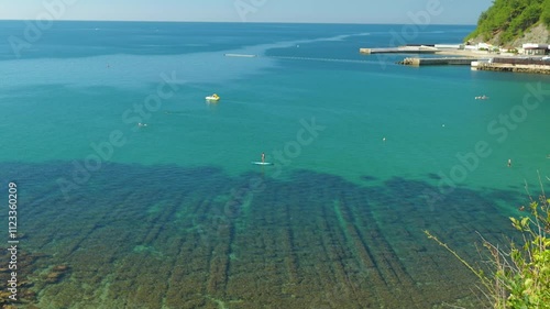 The sea water is crystal clear, with colourful rocks and seaweed visible underneath. People swim and ride m on a paddleboard. Beautiful seascape photo