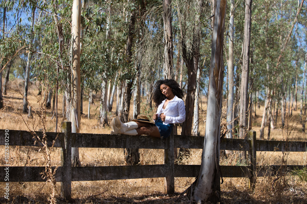 Fototapeta premium woman, young, beautiful, Latin, brunette with curly hair dressed in white shirt, denim skirt, white boots and hat, sitting with her legs stretched out on the forest fence. The woman rests.