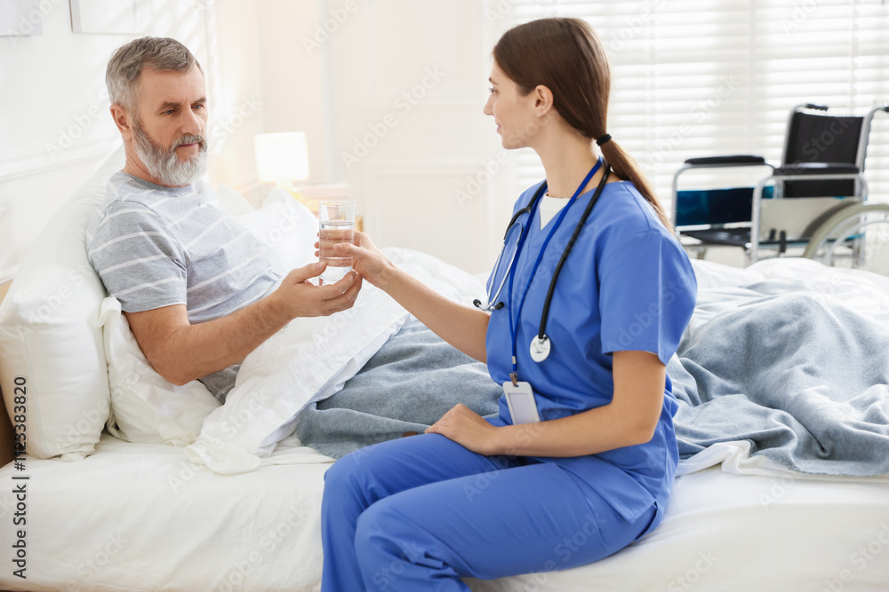 Obraz premium Caregiver giving water to senior man on bed indoors. Home health care service