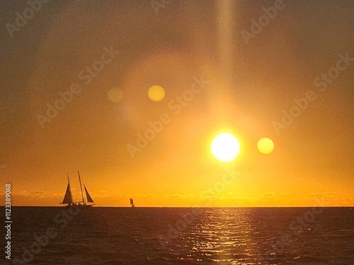 A glowing sun setting over the serene sea, casting warm hues and reflections, with sailing boats dotting the horizon.