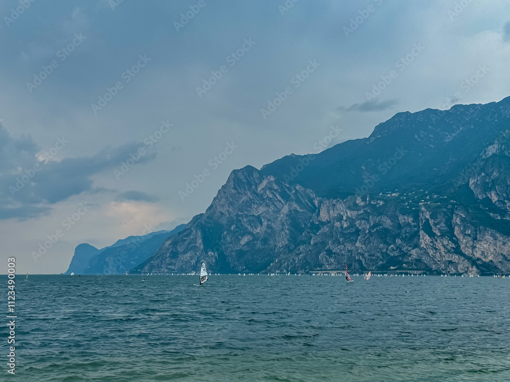 Small sailing boats floating on idyllic Lake Garda surrounded by steep rugged mountains of Garda Prealps. Looking from lakeside town Nago-Torbole, Trentino, Italy. Peaceful summer travel destination Small sailing boats floating on idyllic Lake Garda surrounded by steep rugged mountains of Garda Prealps. Looking from lakeside town Nago-Torbole, Trentino, Italy. Peaceful summer travel destination