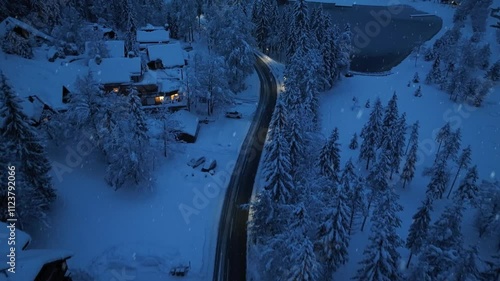 Aerial view of mountain road in fairy forest in snowfall at night. Winter fairytale. Top drone view of mountain village, houses, illumination, snowy pine trees, lake at twilight. Beautiful Slovenia