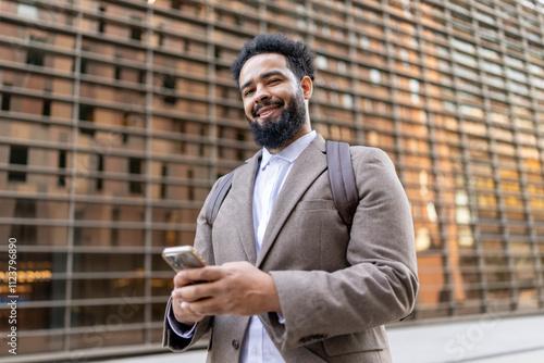 A cheerful African American businessman in a stylish suit confidently stands outdoors by a modern building, holding a smartphone and embracing the vibrant urban environment, reflecting success