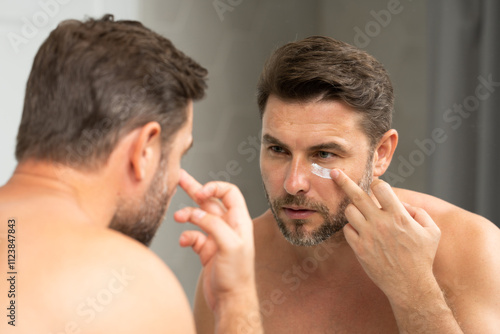 Handsome Man Applying Face Cream in the Bathroom. Closeup beauty portrait of satisfied young man after facial cream. Attractive brunet guy touching skin. Male coscmetic and beauty concept photo