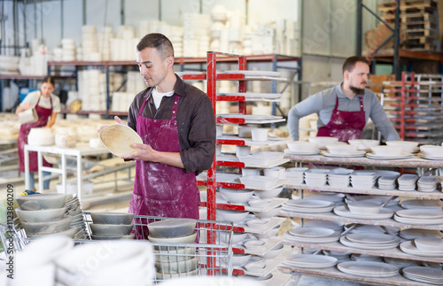 Focused pottery expert in maroon apron meticulously inspecting quality of freshly produced handmade plate in artisanal ceramics studio photo