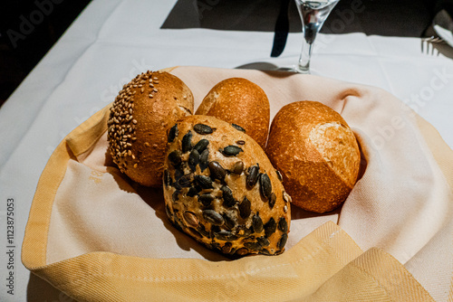 A basket of assorted bread rolls, including sesame seed, plain, and pumpkin seed varieties, served on a cloth napkin in a warm dining setting.