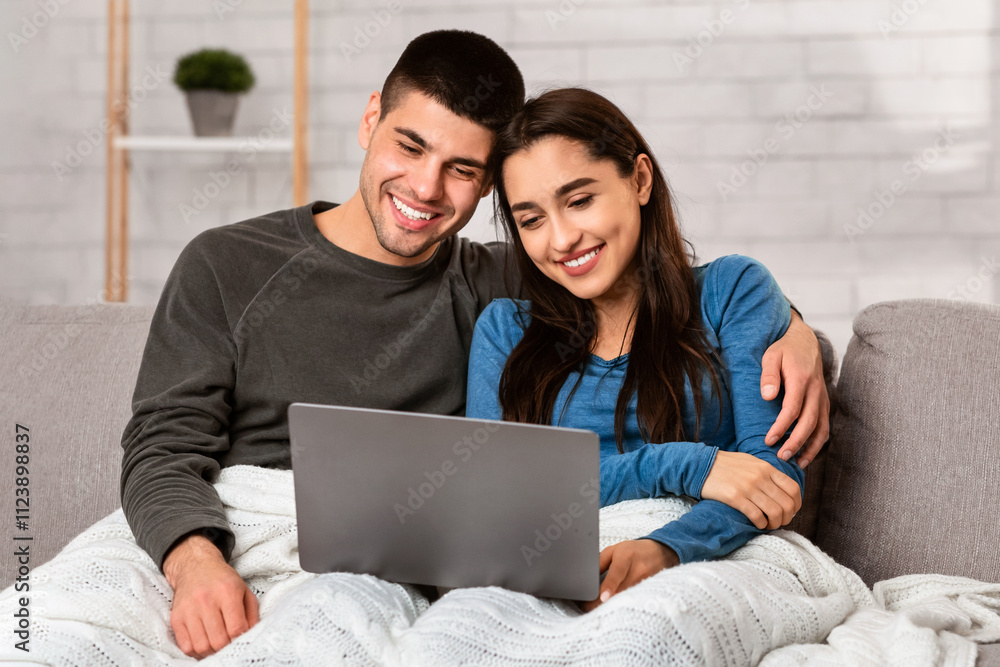 Fototapeta premium Stay At Home Concept. Self isolated spouses hugging and watching movie on laptop sitting on sofa, copy space