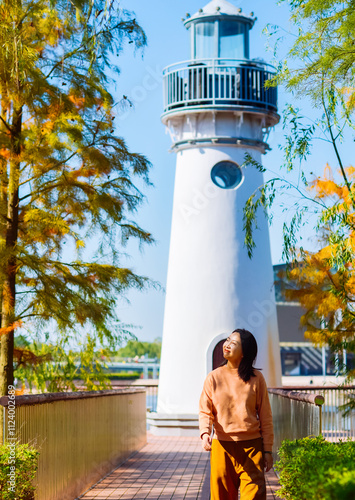 One Asian woman enjoying beautiful landscape of golden autumn season at Yinzhou Park, with white lighthouse and redwood trees in Ningbo, Zhejiang, China