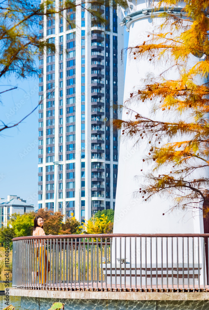 Fototapeta premium One Asian woman enjoying beautiful landscape of golden autumn season at Yinzhou Park, with white lighthouse and redwood trees in Ningbo, Zhejiang, China