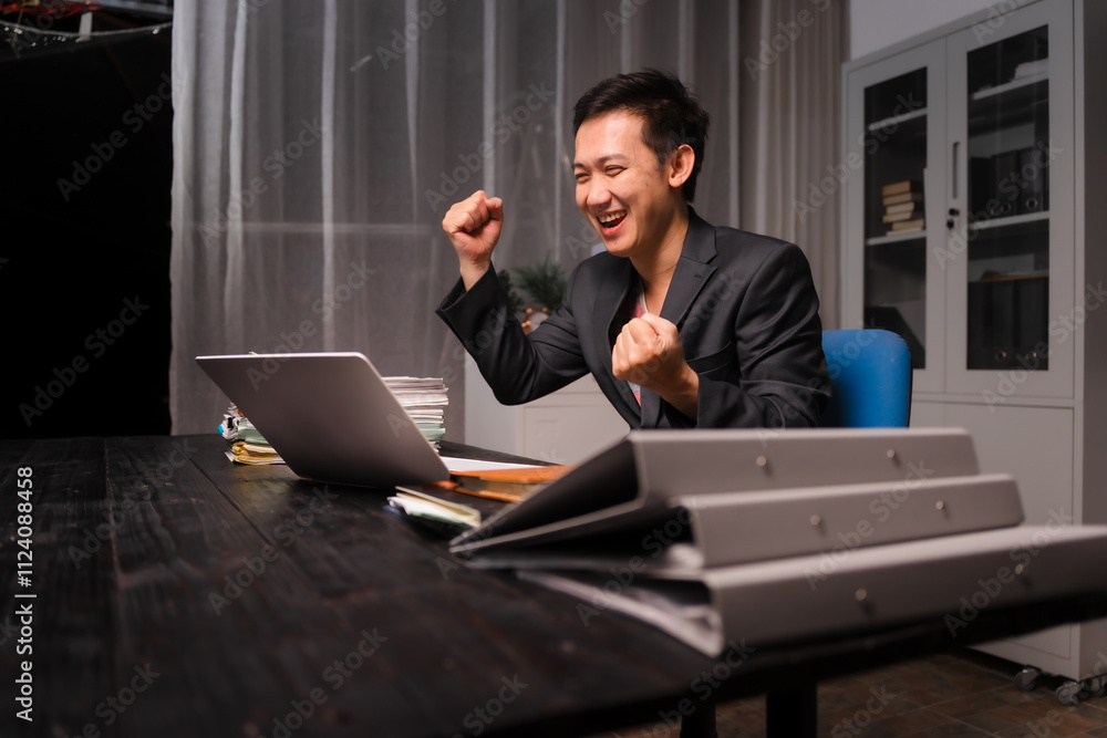 A young Asian man in a formal suit works late at night at his desk, surrounded by paper stacks, analyzing stock market charts on a laptop, celebrating success with a happy smile. A young Asian man in a formal suit works late at night at his desk, surrounded by paper stacks, analyzing stock market charts on a laptop, celebrating success with a happy smile.