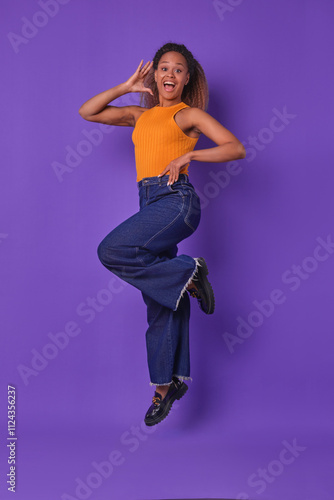 Lively young African American woman is captured mid-jump, wearing a bright orange top and stylish denim jeans. She radiates happiness, showcasing her energetic spirit in a playful moment.