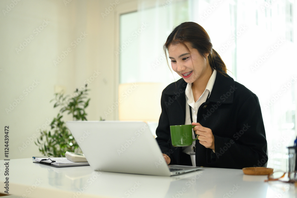 Smiling young businesswoman holding a coffee mug and working on her laptop in office Smiling young businesswoman holding a coffee mug and working on her laptop in office