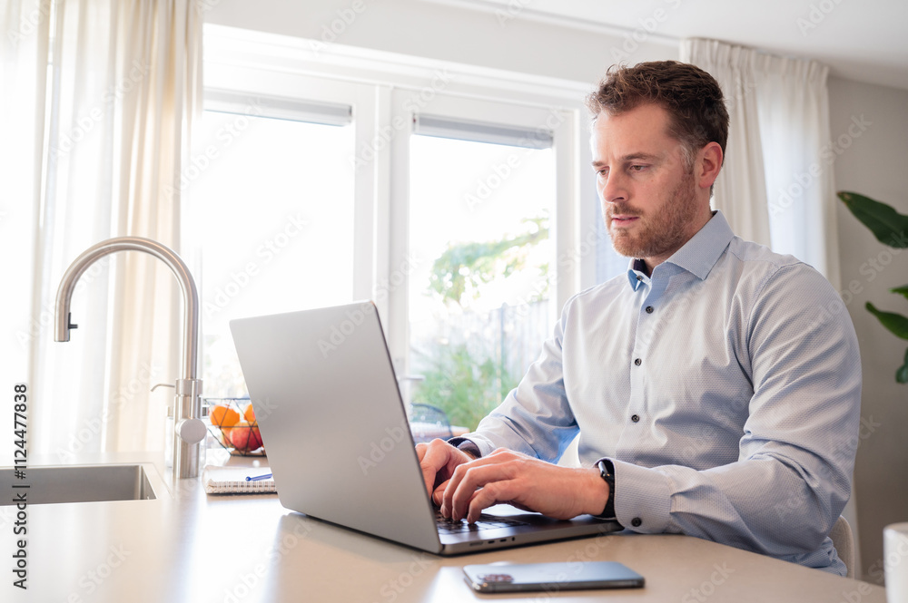 Working at home in the kitchen. Caucasian male sitting at the kitchen counter behind his laptop working and communicating. Wearing a light blue formal shirt