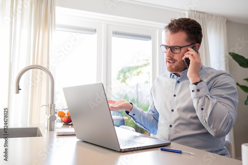 Working at home in the kitchen. Caucasian male sitting at the kitchen counter behind his laptop working and communicating. Wearing a light blue formal shirt and glasses.