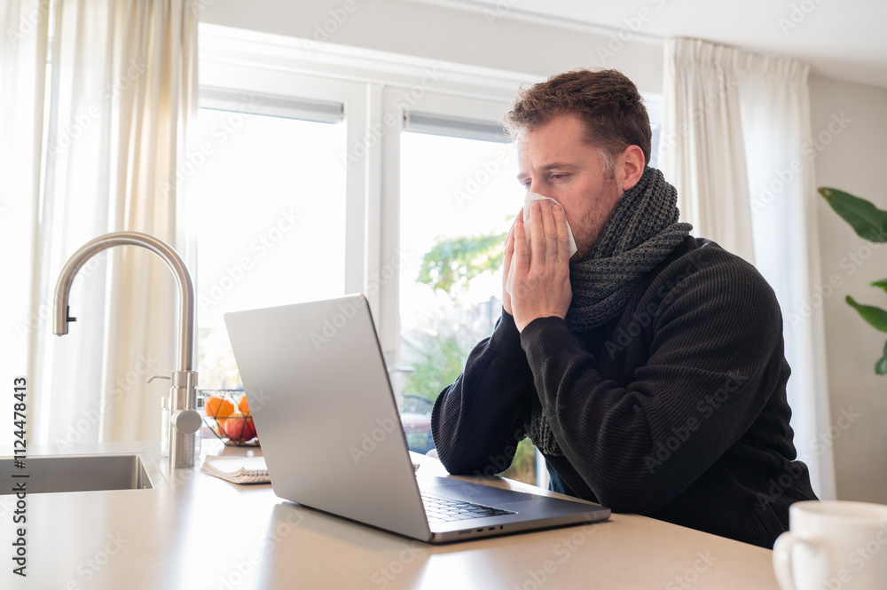 working at home in the kitchen. caucasian male sitting at the kitchen counter behind his laptop working and communicating. Blows his nose in a tissue because he has a cold working at home in the kitchen. caucasian male sitting at the kitchen counter behind his laptop working and communicating. Blows his nose in a tissue because he has a cold