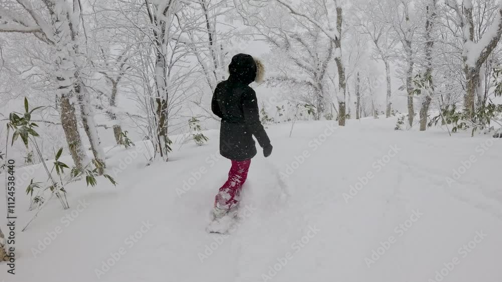 A lone female snowboarder, dressed in black and dark red, makes her way down a secluded forest trail in Rusutsu, Japan, surrounded by snow-heavy trees, showcasing beauty of backcountry snowboarding