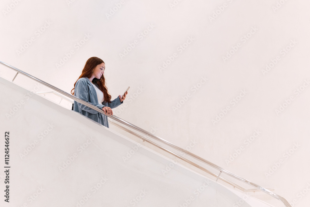 Naklejka premium Side view of young female in stylish wear text messaging on cellphone while standing on stairway in corridor on white background