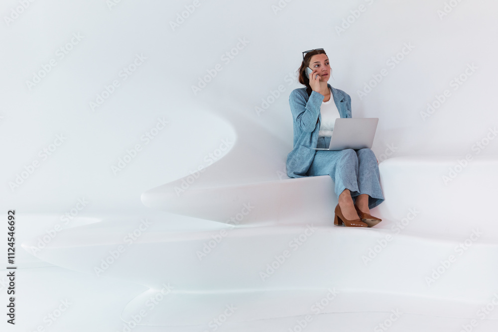 Woman with laptop talking on smartphone. Full body of elegant female in stylish wear browsing laptop while having phone conversation in spacious light studio with white wall Woman with laptop talking on smartphone. Full body of elegant female in stylish wear browsing laptop while having phone conversation in spacious light studio with white wall