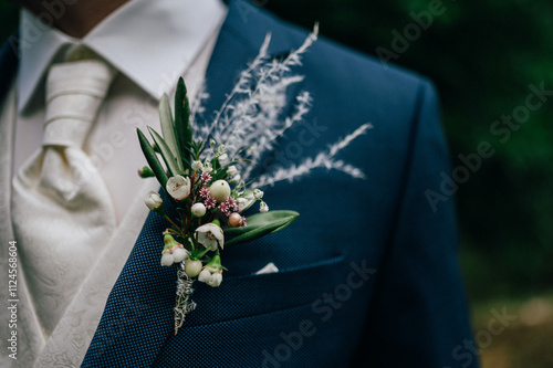 Nahaufnahme eines Bräutigams in einem dunkelblauen Anzug mit einer eleganten Ansteckblume am Revers. Die Blume besteht aus weißen Beeren, grünen Blättern und feinen Gräsern.