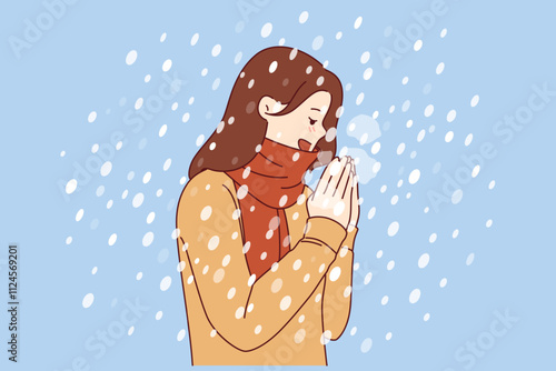 Woman enjoys winter weather, stands under falling snow flakes in sweater and scarf. Positive girl rejoices at onset of winter and approach of christmas holidays, blowing on hands to keep warm