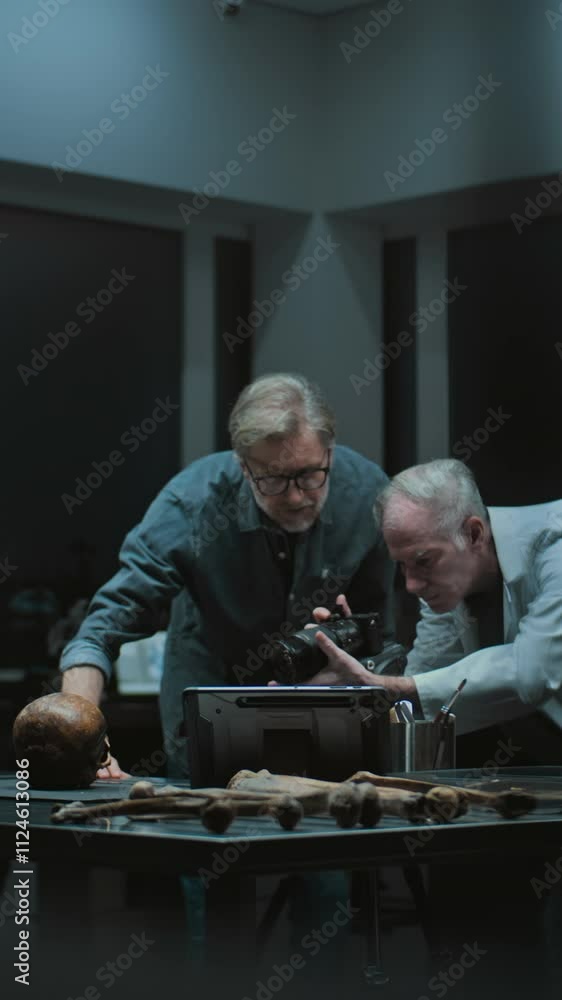 Two archaeologists take picture of fossil remains of prehistoric human skull with professional camera on tripod. Scientists work in modern archaeological laboratory. Vertical shot Two archaeologists take picture of fossil remains of prehistoric human skull with professional camera on tripod. Scientists work in modern archaeological laboratory. Vertical shot