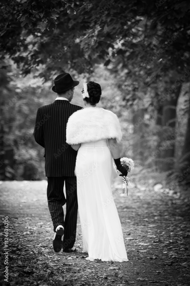 A man and woman are walking together in a forest, dressed in black and white attire. The man is wearing a hat and the woman is wearing a fur coat. They are holding hands. A man and woman are walking together in a forest, dressed in black and white attire. The man is wearing a hat and the woman is wearing a fur coat. They are holding hands.
