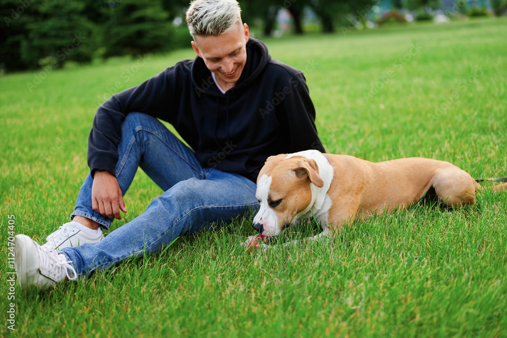 Dog lying on the grass playing with a ball, the owner is sitting next to him and stroking the dog. Love to the dog as a member of the family. American bulldog. Dog lying on the grass playing with a ball, the owner is sitting next to him and stroking the dog. Love to the dog as a member of the family. American bulldog.