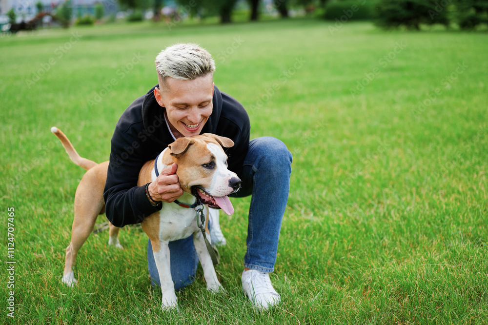 Dog walker bending down to stroke a dog nuzzling his face. A young man fooling around with his dog in the park. Man's best friend. Man and american bulldog. Dog walker bending down to stroke a dog nuzzling his face. A young man fooling around with his dog in the park. Man's best friend. Man and american bulldog.