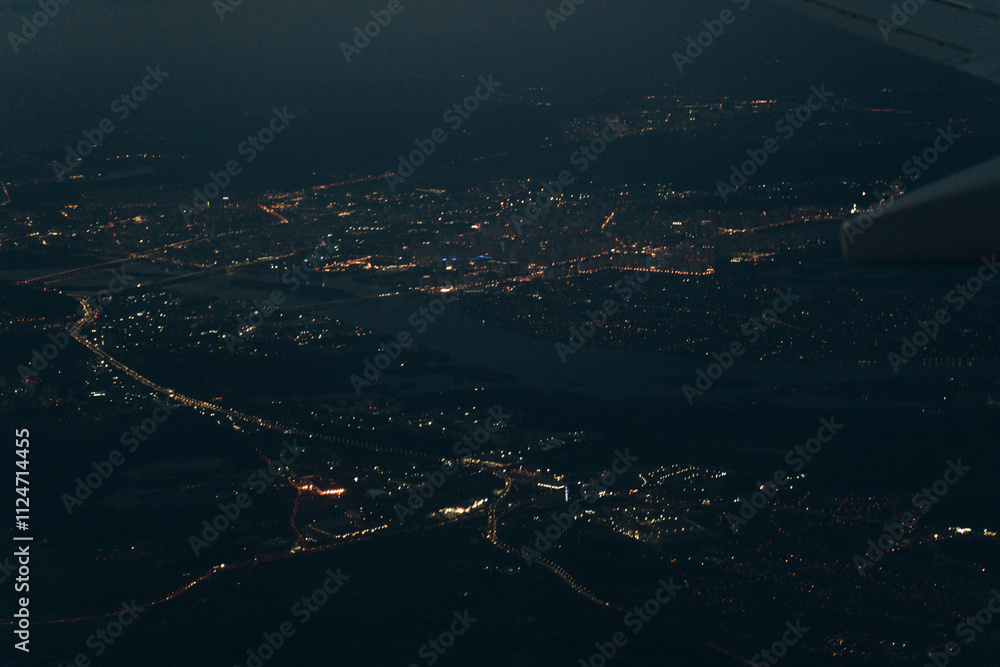 Fototapeta premium Aerial view from airplane window of buildings and bright illuminated streets in city residential area at night. Dark urban landscape at high altitude