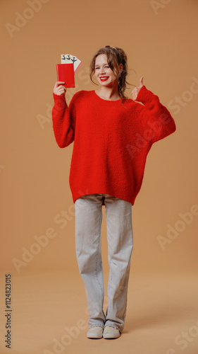 Cheerful young woman in red jumper and grey jeans posing with flight tickets, smiling shows thumb up like gesture, isolated on orange studio background photo