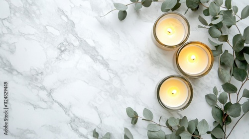 Overhead view of candles in glass holders with eucalyptus on a white marble background with space for text