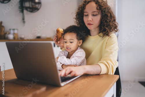 Working mother balancing professional life and parenting in home kitchen interior. Mixed-race child enjoys morning snack during casual mommy business meeting via modern laptop computer connected to 4g