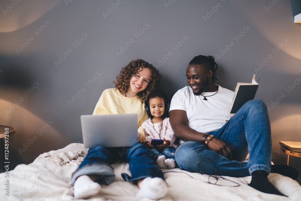 Cheerful mom using laptop technology while father smiling at camera during relaxed moment with their pretty child in headphones,portrait of happy marriage with daughter spending multitasking weekend Cheerful mom using laptop technology while father smiling at camera during relaxed moment with their pretty child in headphones,portrait of happy marriage with daughter spending multitasking weekend