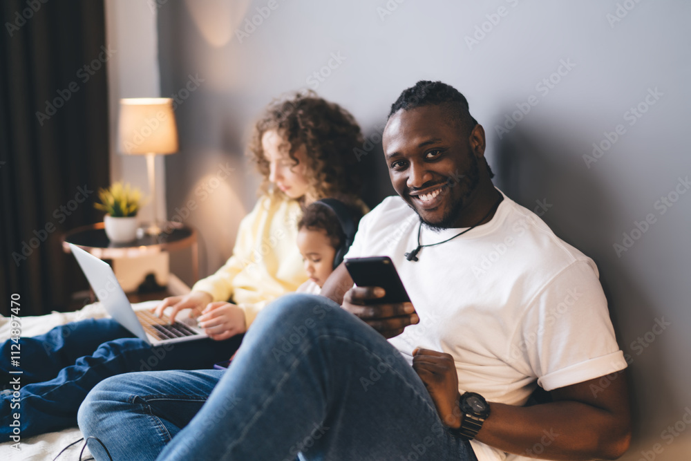 Portrait of cheerful dark skinned male with modern cellphone technology smiling at camera during weekend time with blurred family, happy African American hipster guy with smartphone device posing Portrait of cheerful dark skinned male with modern cellphone technology smiling at camera during weekend time with blurred family, happy African American hipster guy with smartphone device posing