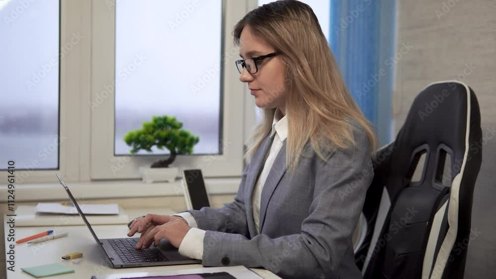caucasian thoughtful female in glasses sitting at the table using notebook. Education and business concept. A business girl is working at a laptop in a white fleece. caucasian thoughtful female in glasses sitting at the table using notebook. Education and business concept. A business girl is working at a laptop in a white fleece.