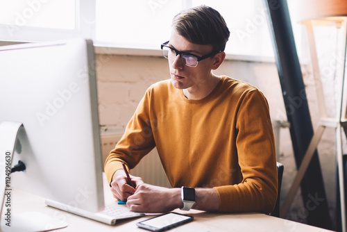 Young hipster guy in spectacles watching important webinar on web page internet via modern computer while writing main information, caucasian man reading news in social network via technology photo