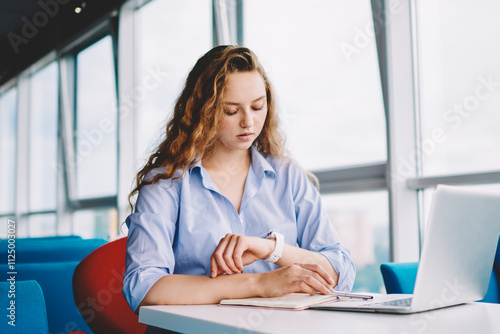 Serious female freelancer checking notification on digital smartwatch while working distance in cafeteria on laptop device, concentrated woman reading organiser notes on electronic wristwatch