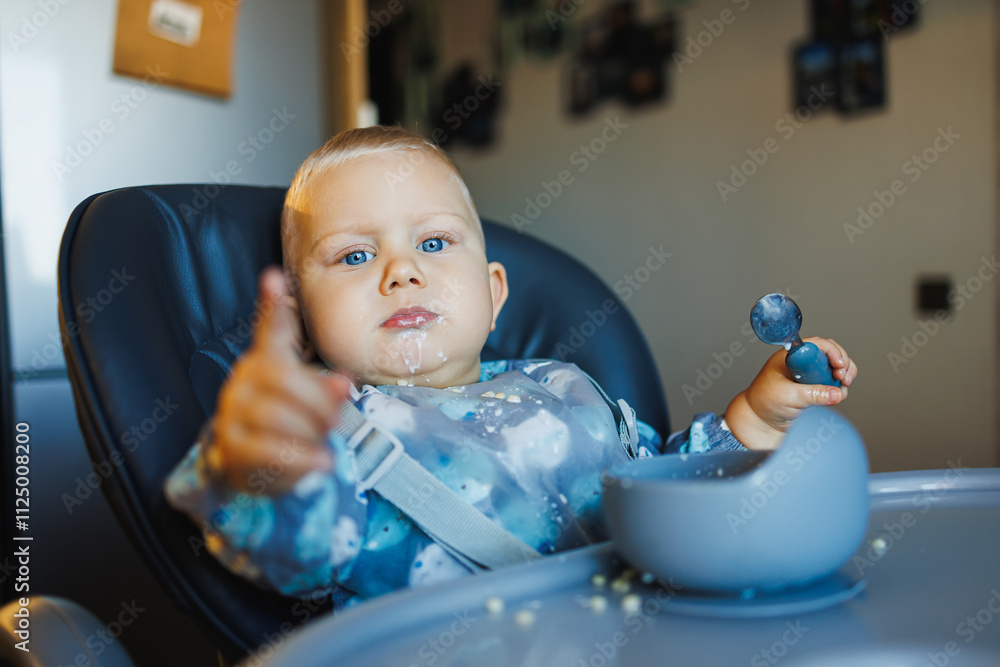 Cute one-year-old boy sits in a high chair and eats independently. Silicone utensils for feeding children. Cute one-year-old boy sits in a high chair and eats independently. Silicone utensils for feeding children.