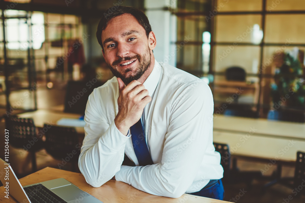 Fototapeta premium Portrait of cheerful male economist in formal wear sitting at desktop with laptop device and smartphone and looking at camera, successful man employee spending time in office and waiting colleague
