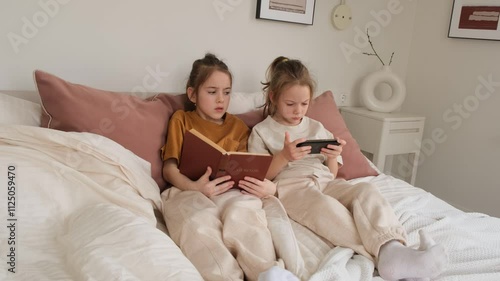 Two twin sisters relax in bed, one engrossed in a book and the other focused on her smartphone. A peaceful, intimate moment capturing sibling interaction and quiet leisure time at home.