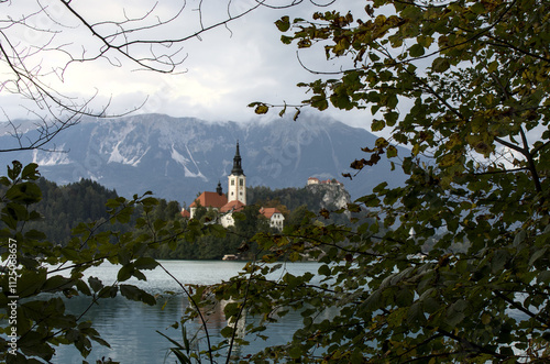 view of the island on lake bled through beautiful autumn tree leaves (foliage on famous travel destination landmark) julian alps slovenia europe european destination fairytale setting foreground photo