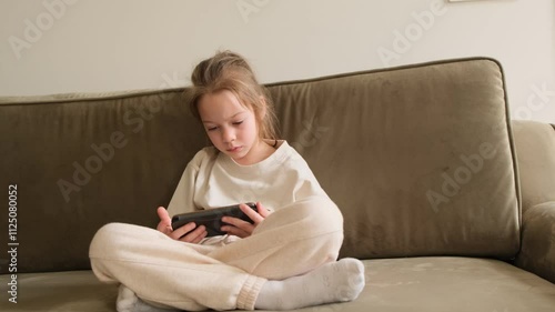 A young girl sits on a couch, engrossed in a smartphone. The focus is on her and the neutral colors of the couch and her clothing. The scene suggests a relaxed, quiet moment at home.
