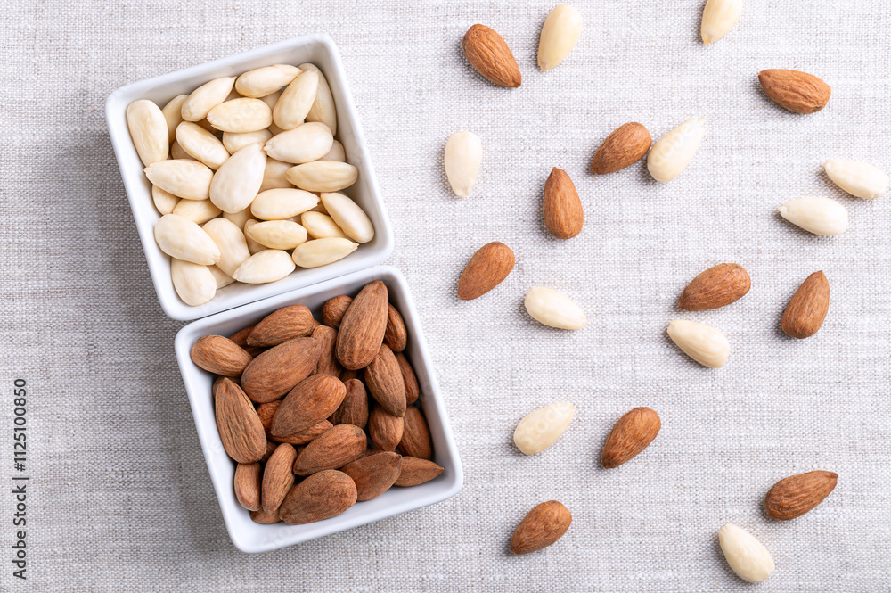 Almonds in square shaped white bowls, on linen fabric. Almonds with seed coats and blanched. Fruits of Prunus amygdalus, syn. Prunus dulcis, ready-to-eat as snack or as ingredient for baking. Photo.