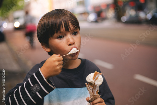 Little brunette boy eat ice cream in waffle corn with spoon. Happy childhood. Boy walk outdoor in the city street. 