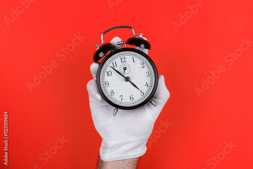 A white-gloved hand holds a classic black alarm clock against a bold red background, highlighting time management and punctuality. Near five o clock photo