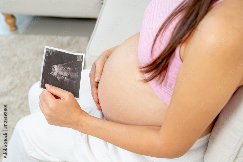 Side and high view of pregnant woman use left hand to hold ultrasound image of her baby and sit on sofa in living room. Side and high view of pregnant woman use left hand to hold ultrasound image of her baby and sit on sofa in living room.