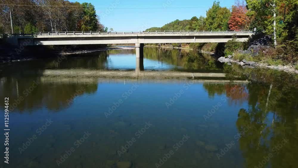 Flight over a calm and clear river which reflects the surrounding scenery while passing under a reinforced concrete pillar bridge in an autumnal setting. Flight over a calm and clear river which reflects the surrounding scenery while passing under a reinforced concrete pillar bridge in an autumnal setting.