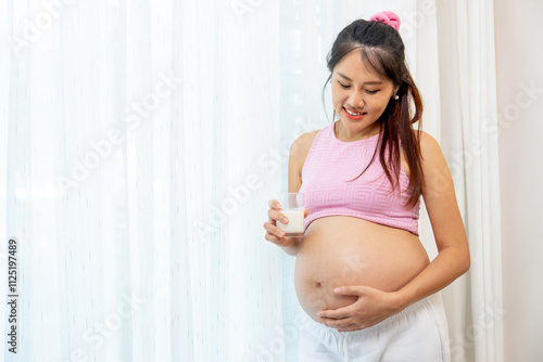 Asian pregnant woman stand with hold a glass of milk and stand in front of white curtain also look at her belly with happiness smile. Free Download Asian pregnant woman stand with hold a glass of milk and stand in front of white curtain also look at her belly with happiness smile. - GFXLoad