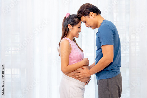 Asian man and pregnant woman stand with hug and attach forehead together express love and care in front of white curtain.